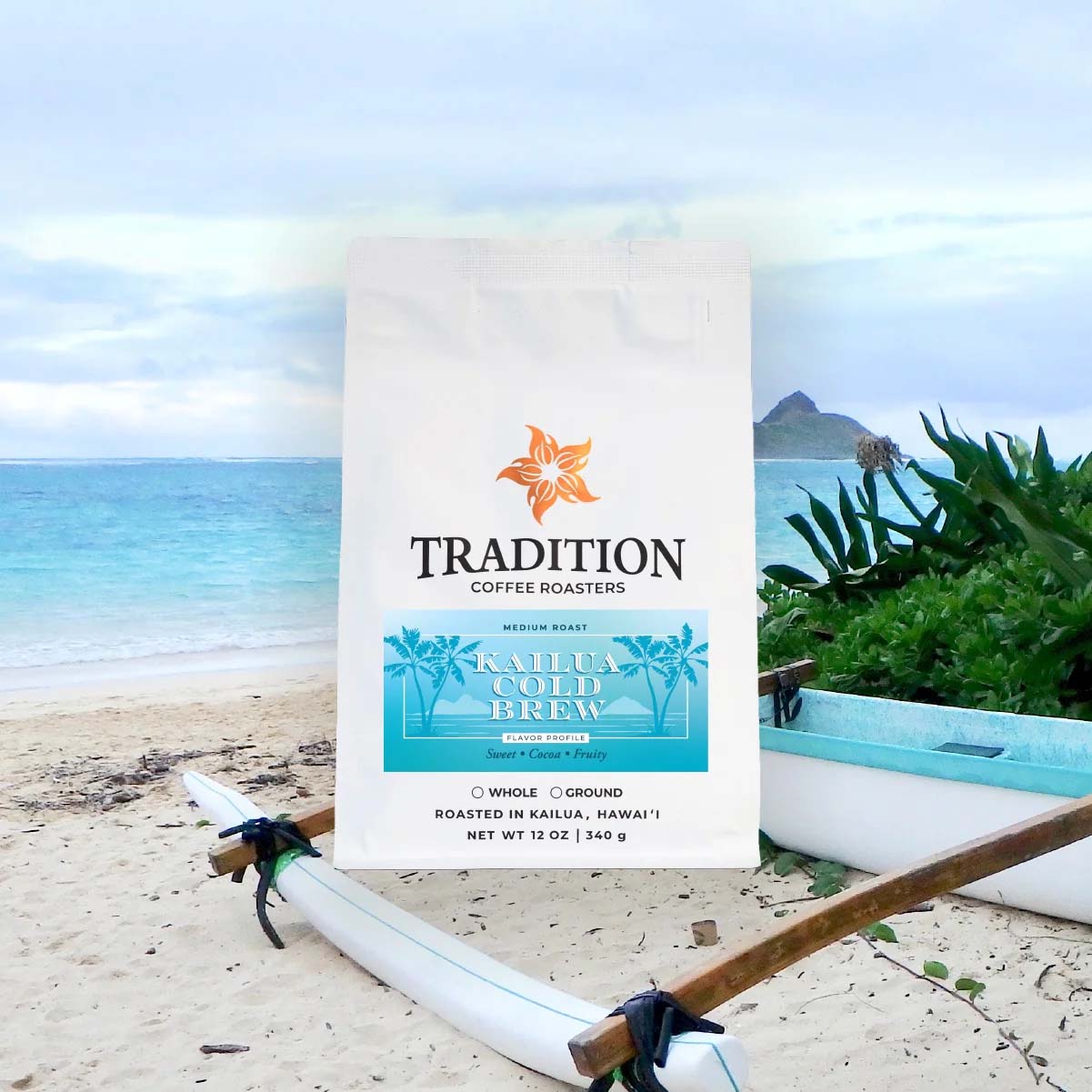 A bag of Tradition Coffee Roasters Kahua Cold Brew coffee stands on a sandy beach with a surfboard, tropical plants, and the ocean in the background
